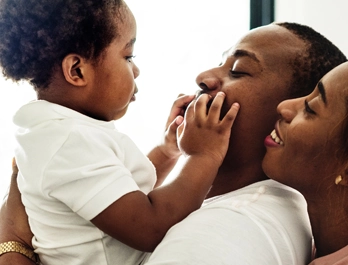 A father holds a young child in his arms, smiling as the child feels and explores his face. Mom stands behind Dad, smiling at the child.