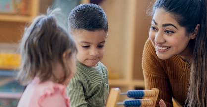 A woman smiles at two toddlers, a girl and a boy, playing with an abacus-style toy.