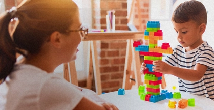 A woman watches on as a small boy builds a colorful structure with interlocking blocks on a table.