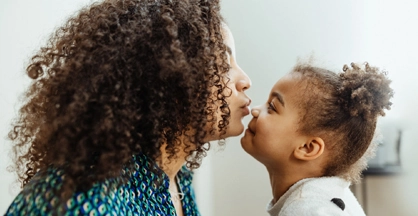 A woman and a small child face each other. The woman is poised to kiss the tip of the little girl's nose while the girl smiles.