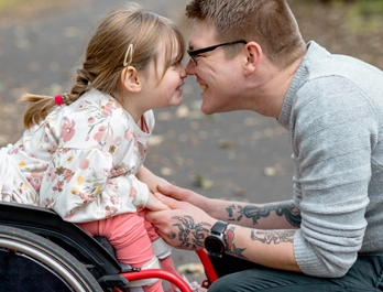 A young girl, seating in a wheelchair, smiles as she touches noses with her father. The father holds her hands and smiles as he kneels in a leaf-strewn path.