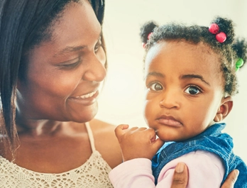 A little girl with colorful hair ties looks at the camera, held by a woman who smiles at her.