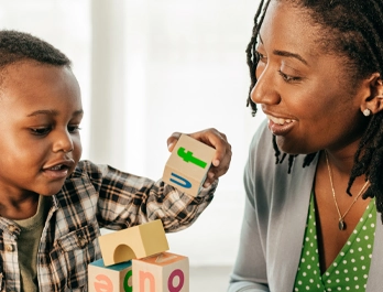 A little boy builds with blocks while a woman watches encouragingly.