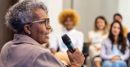 A woman with gray hair and glasses holds a microphone and speaks to an audience.