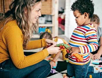 A woman and a little boy kneel together on the floor, holding connector blocks with wheels.