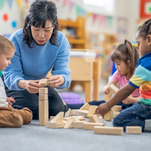 A woman kneels on the ground, building blocks with three toddlers.