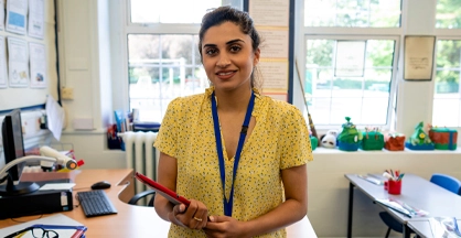 A woman wearing a lanyard stands in a classroom, smiling and holding a folder.