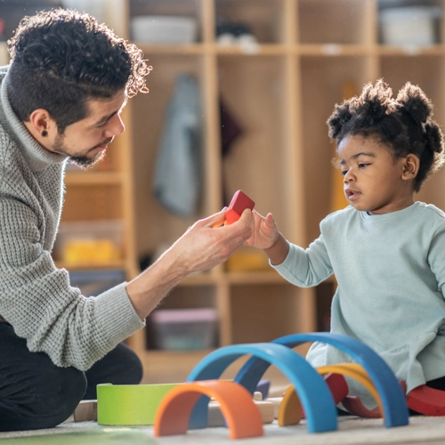 A man and toddler are seated on the ground, playing with shapes. The man hands a block to the toddler.