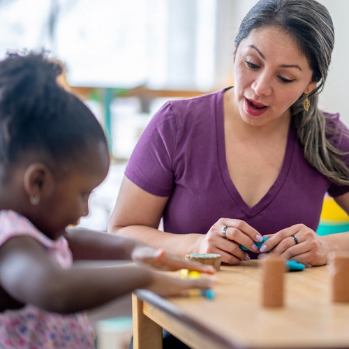 A woman and young girl sit together at a table, playing, while the woman speaks encouragingly