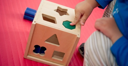 A child's hand pushes an octagonal shape through an octagonal hole in a wooden square.