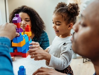 A man and woman help a smiling little girl build a tower of blocks