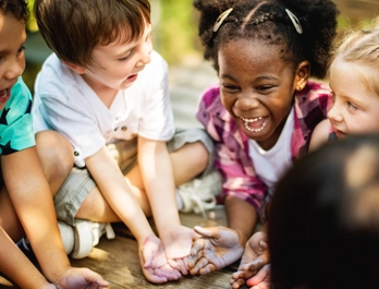 A group of children are seated in a circle outside, as a young girl laughs