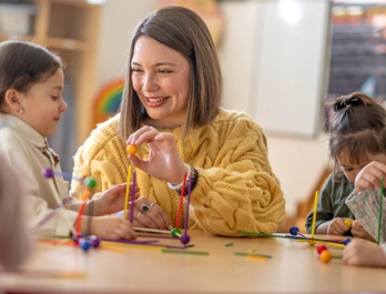 A woman smiles at a little girls as the build structures together at a table with other children.