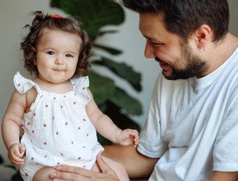 A smiling toddler looks to the camera as a man holds her and smiles.