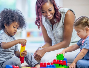 A woman kneels on the floor and smiles as two toddler stack blocks