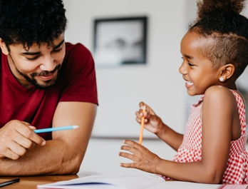 A man and young girl are seated at a table, holding pencils and talking
