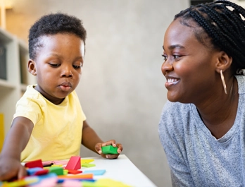 A woman smiles at a toddler playing with blocks at a table.