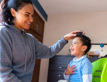 A woman and young boy smile at each other as she arranges his hair.