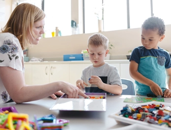 A woman draws attendtion to colorful items in a box as two young boys look on in a classroom.