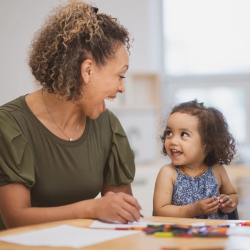 A woman and toddler girl are seated at a table with coloring implements, looking at each other with big smiles.