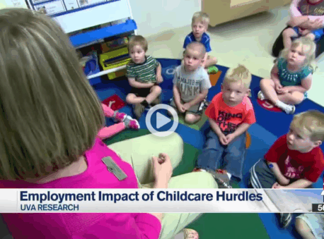 A group of toddlers sits on the floor, listening to a teacher read to them.