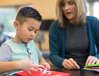 A woman encourages a little boy as he works on a project at a table.