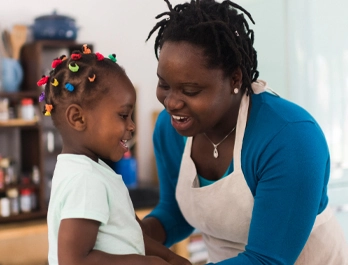 A woman with an apron reaches out to a little girl with colorful hair ties as both smile.