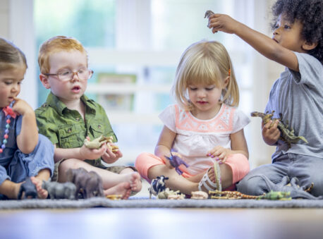 A small group of toddlers sit on the carpet in a home daycare as they play together with toy animals. They are each dressed casually as they share and play together quietly.