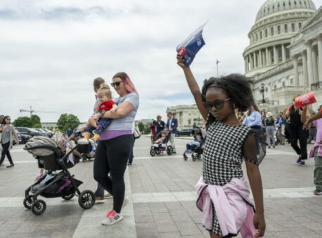A little girl holds up a flyer in front of the US capitol building, walking with other families and children.